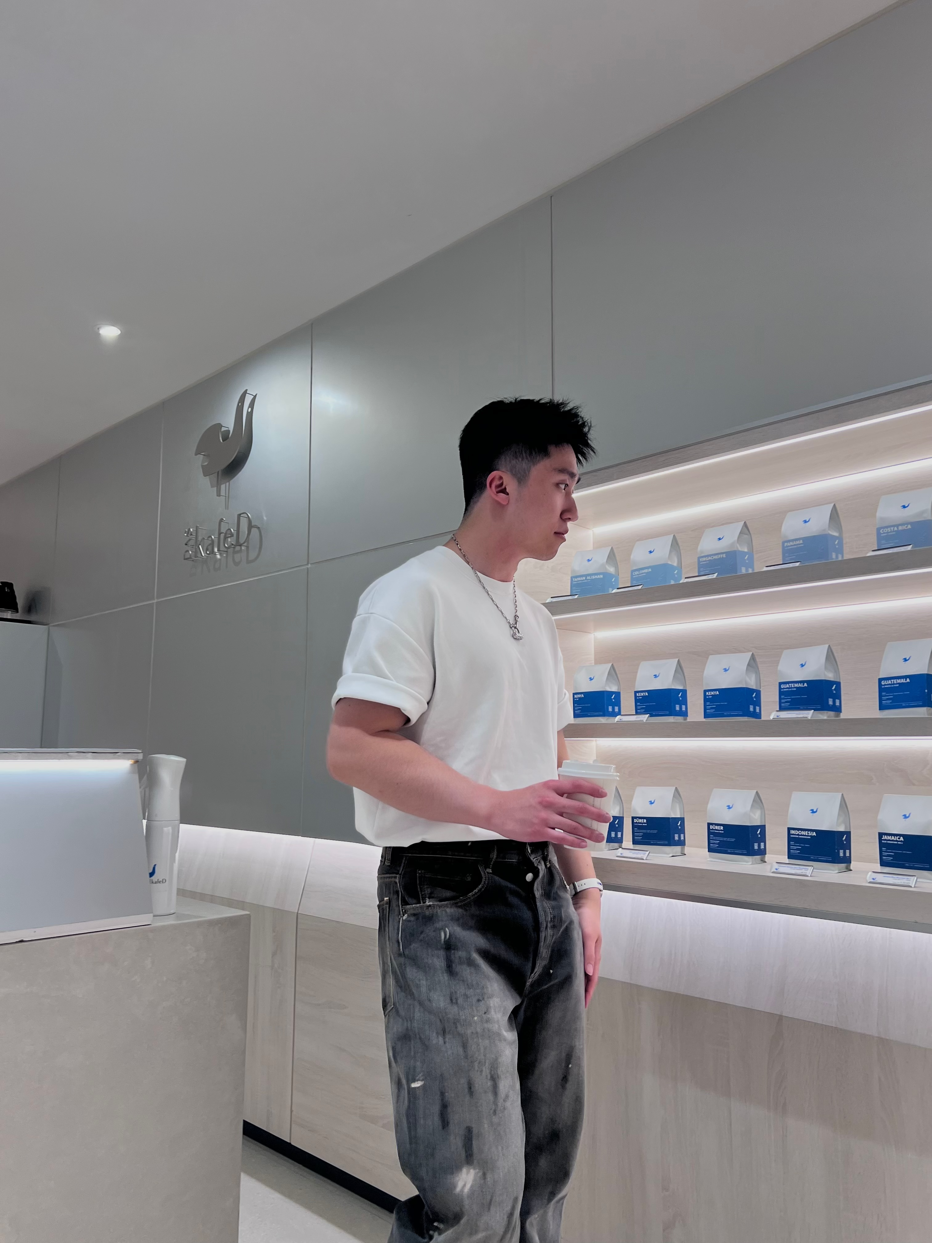Chenwei Lin standing in a minimal café interior, holding a coffee cup beside shelves of coffee bags